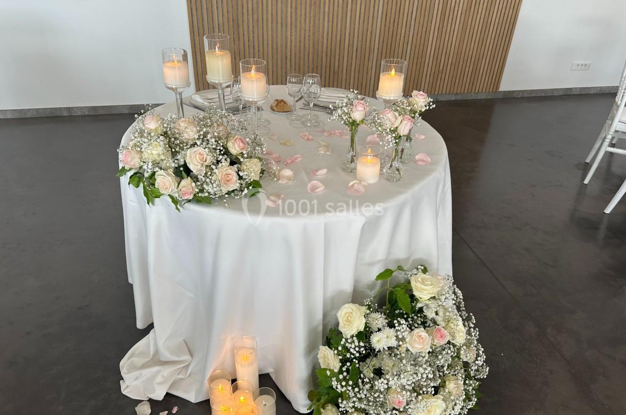 Table ronde décorée de bougies, pétales de rose et compositions florales blanches et roses, dans une salle sobre.