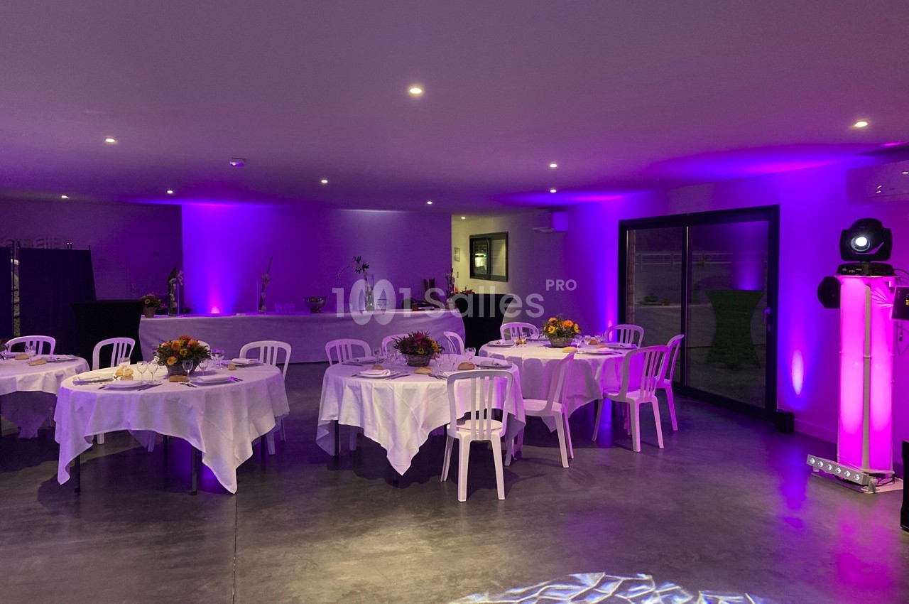 Salle de réception décorée avec des tables rondes, nappes blanches, chaises et éclairage violet tamisé.