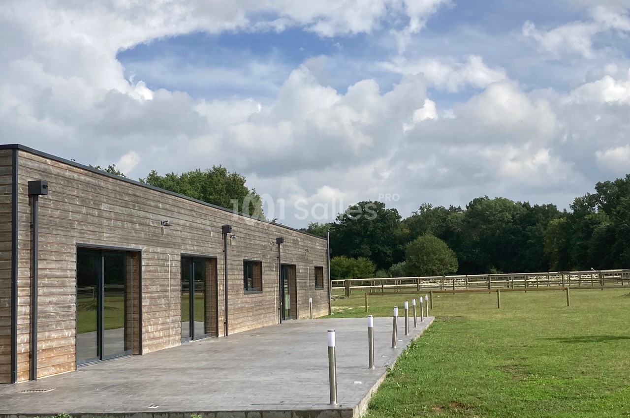 Bâtiment en bois avec une terrasse en béton, entouré de pelouses et de clôtures dans un environnement rural.