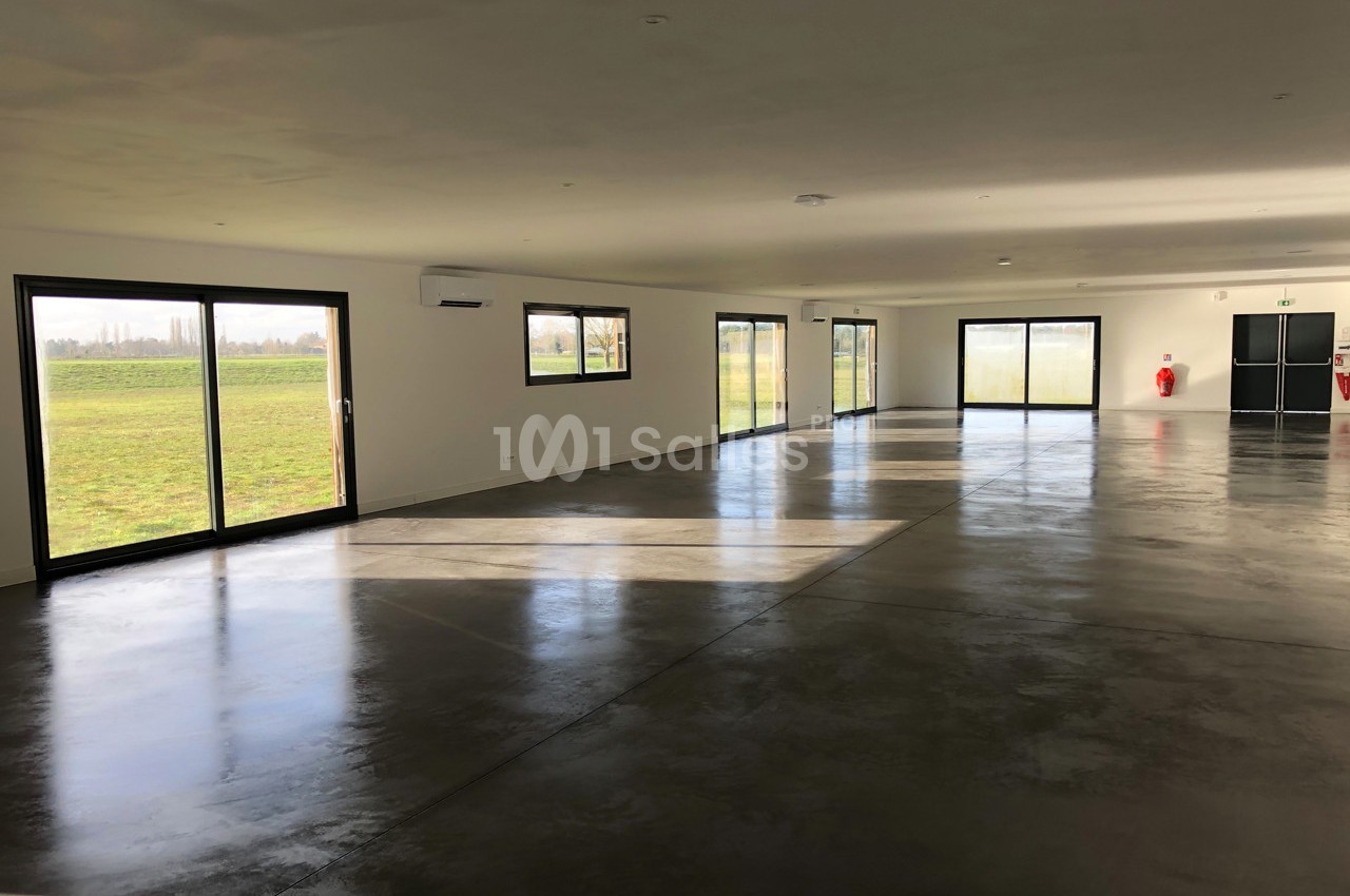 Salle vide avec sol en béton poli, grandes baies vitrées laissant entrer la lumière naturelle et vue sur un espace vert.