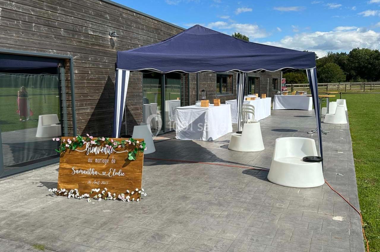 Table et chaises blanches sous un barnum sur une terrasse pavée, avec une pancarte en bois décorée au premier plan.