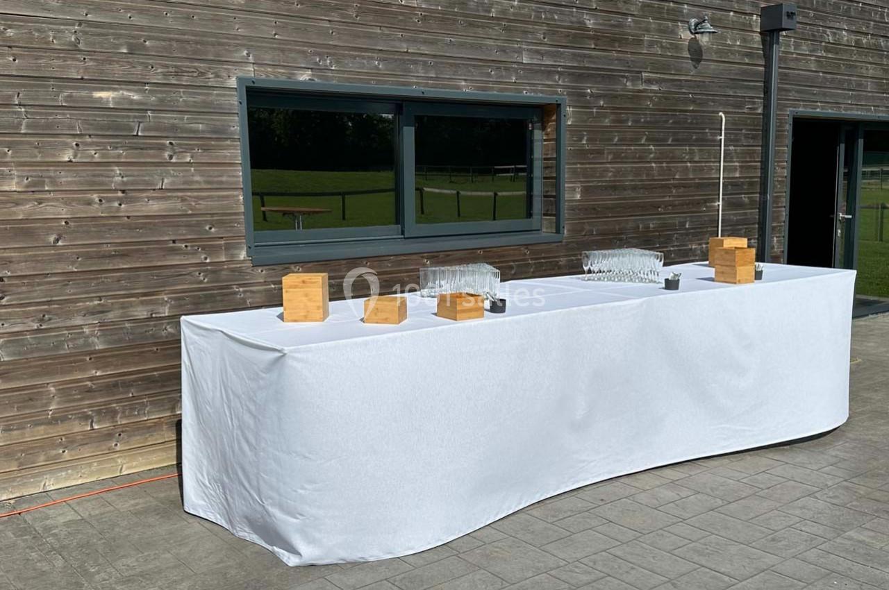 Table recouverte d'une nappe blanche, disposée devant un mur en bois, avec des verres et des boîtes en bois.