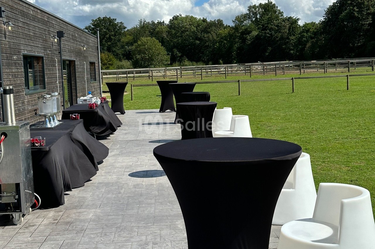 Tables hautes noires et chaises blanches disposées sur une terrasse ensoleillée près d'une pelouse et d'une clôture en bois.