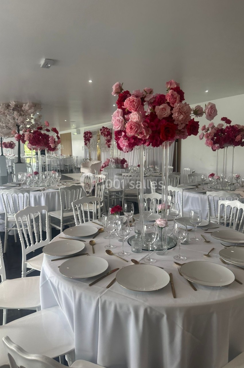 Salle de réception décorée avec des tables rondes, nappes blanches et centres de table composés de fleurs roses.