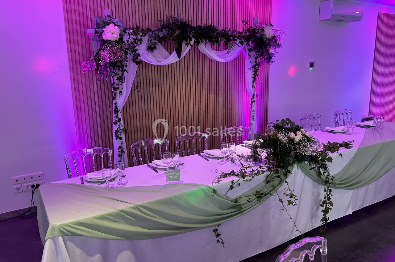 Table de réception décorée avec des nappes blanches, des fleurs, des feuillages et un arrière-plan éclairé en rose.
