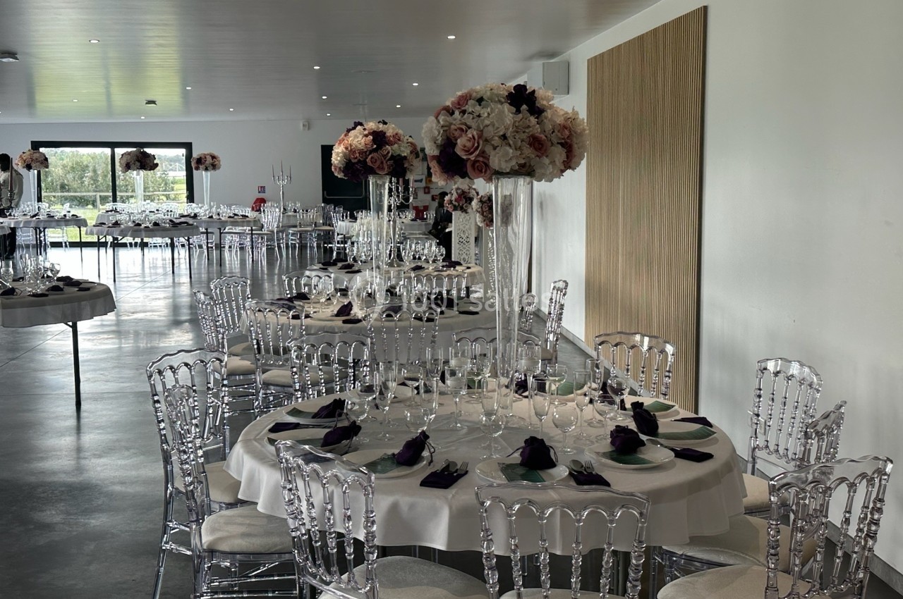 Salle de réception élégamment décorée avec des tables rondes, nappes blanches, chaises transparentes et centres de table…