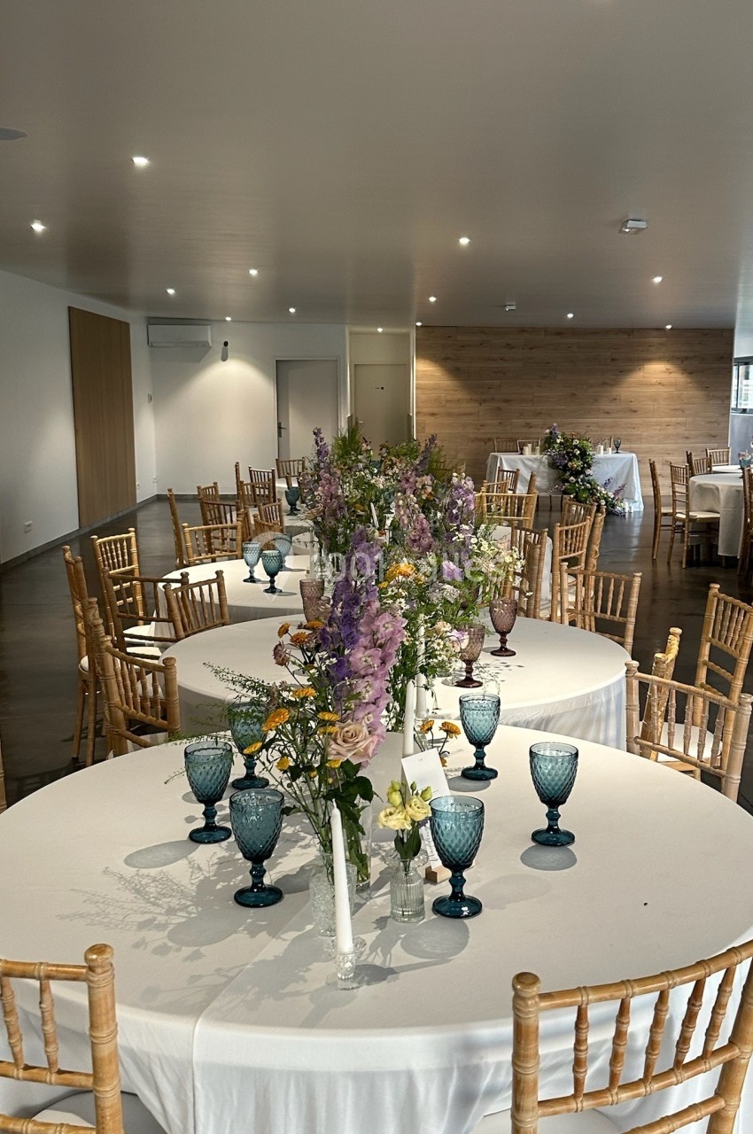 Salle de réception décorée avec des tables rondes, nappes blanches, chaises en bois et bouquets de fleurs.