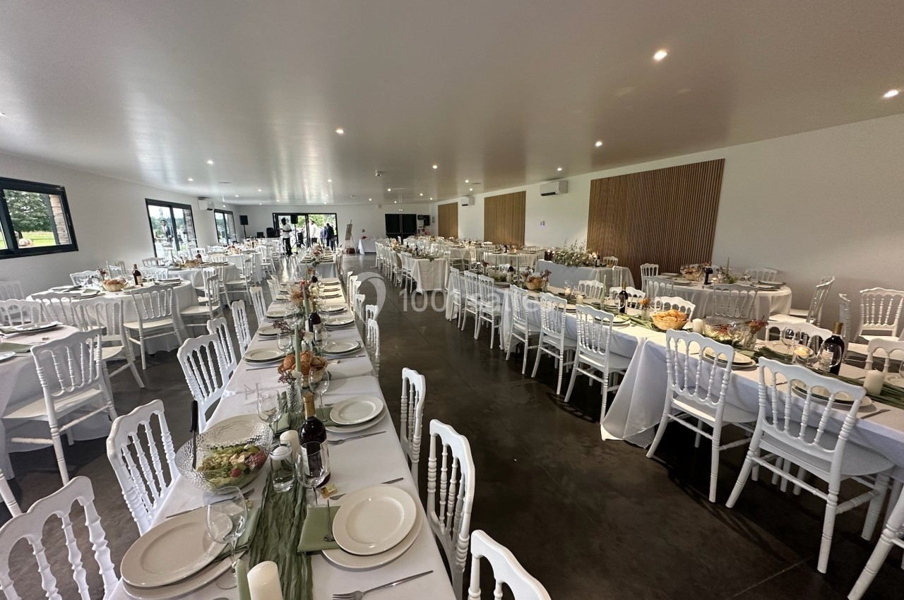 Salle de réception décorée avec des tables alignées, nappes blanches, chaises en bois et centre de table floral.