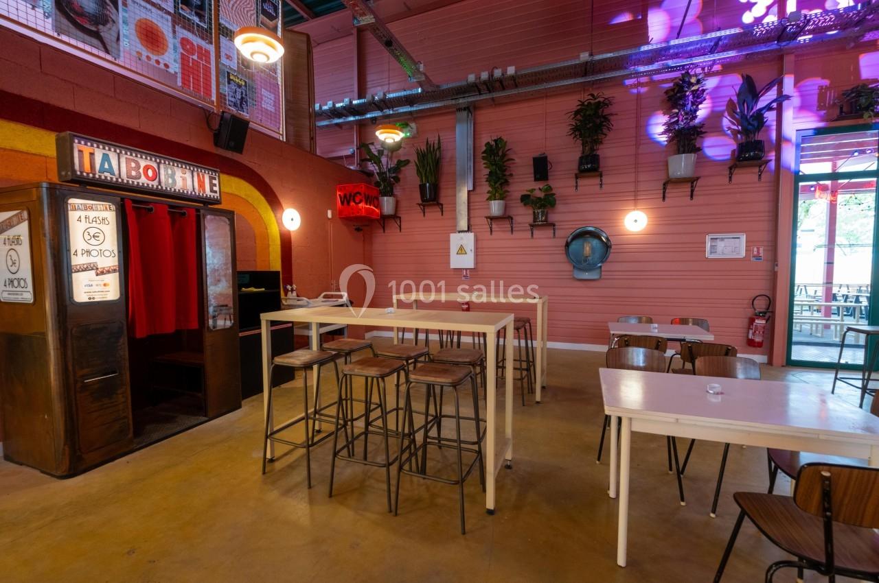 Salle lumineuse avec tables hautes, chaises, plantes murales et un photomaton rétro dans un décor industriel.