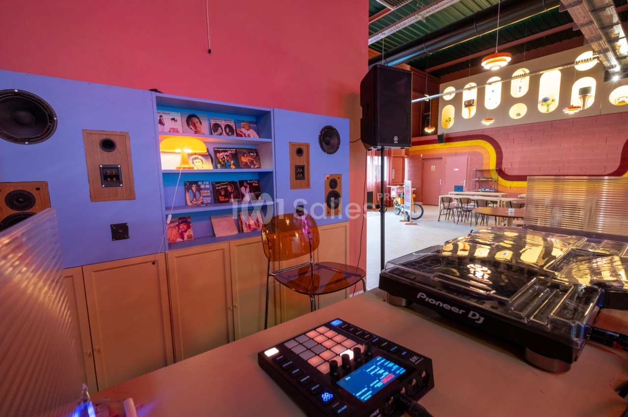 Salle colorée avec platine DJ, enceintes murales, étagère de vinyles et mobilier rétro dans un espace lumineux.