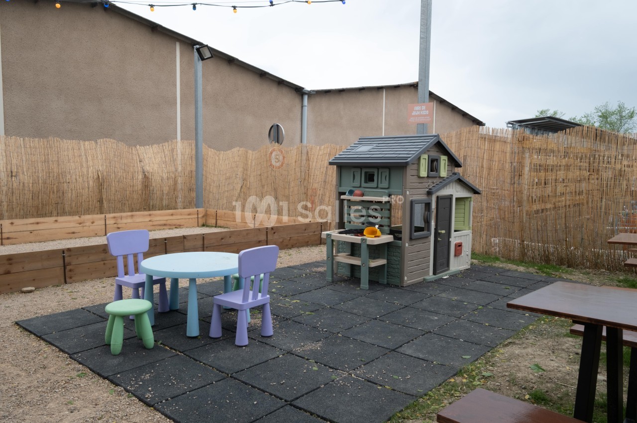 Espace de jeu extérieur avec une maisonnette en plastique, une table ronde et des chaises colorées sur un sol en dalles…