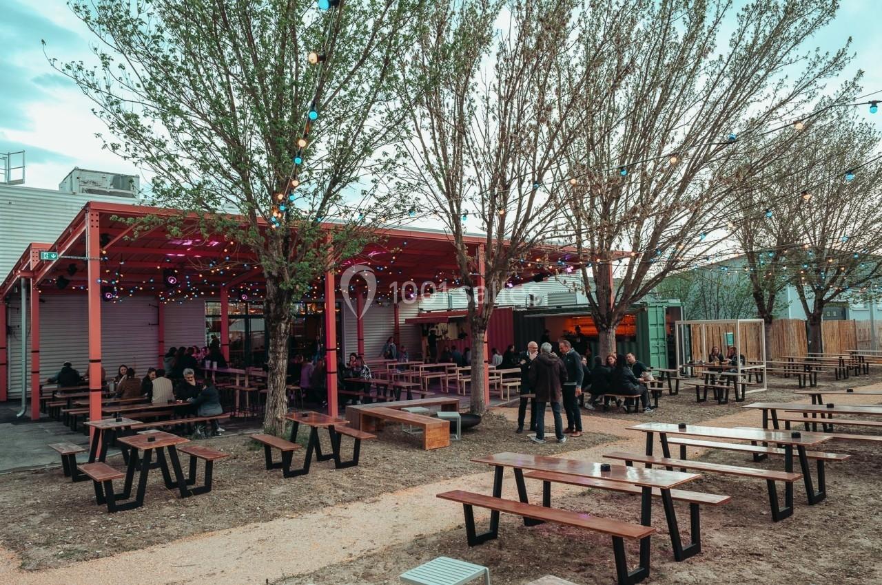 Terrasse extérieure avec tables en bois, éclairages suspendus et quelques personnes assises sous un auvent rouge.