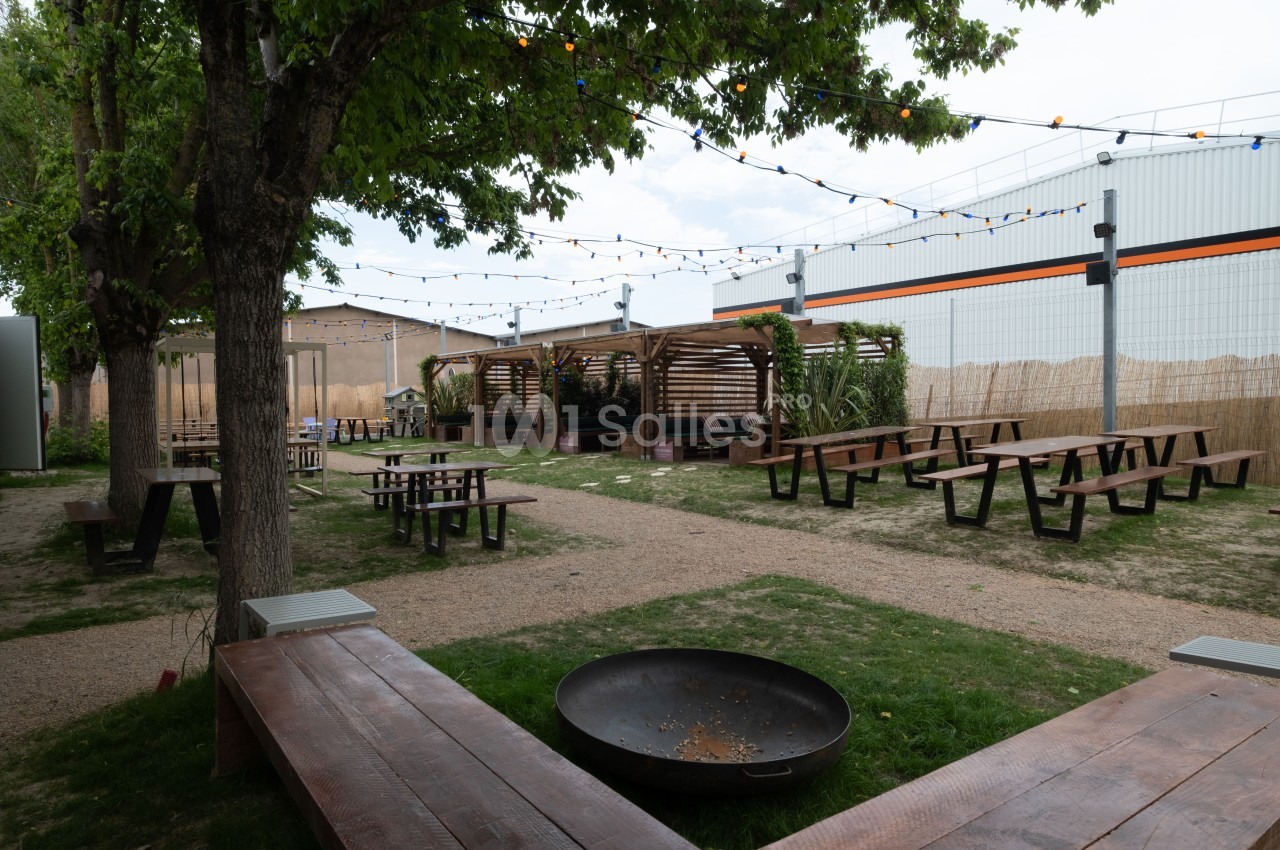 Espace extérieur avec tables en bois, bancs, arbres, guirlandes lumineuses et brasero sur une pelouse.