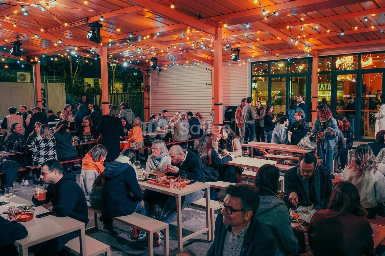 Terrasse animée en soirée avec des tables occupées par des personnes partageant un repas sous des guirlandes lumineuses.