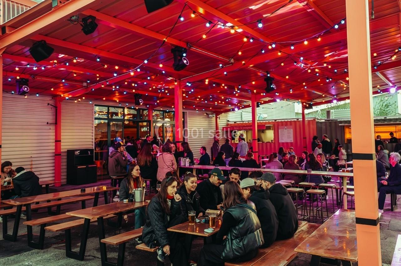 Terrasse extérieure animée avec des tables en bois, des lumières colorées suspendues et des groupes de personnes discutant.