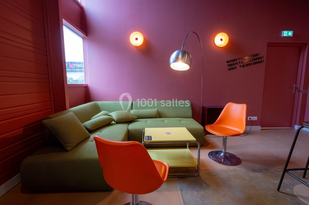 Salon moderne avec canapé vert, deux chaises orange, table basse jaune et lampadaire métallique dans une pièce aux murs…