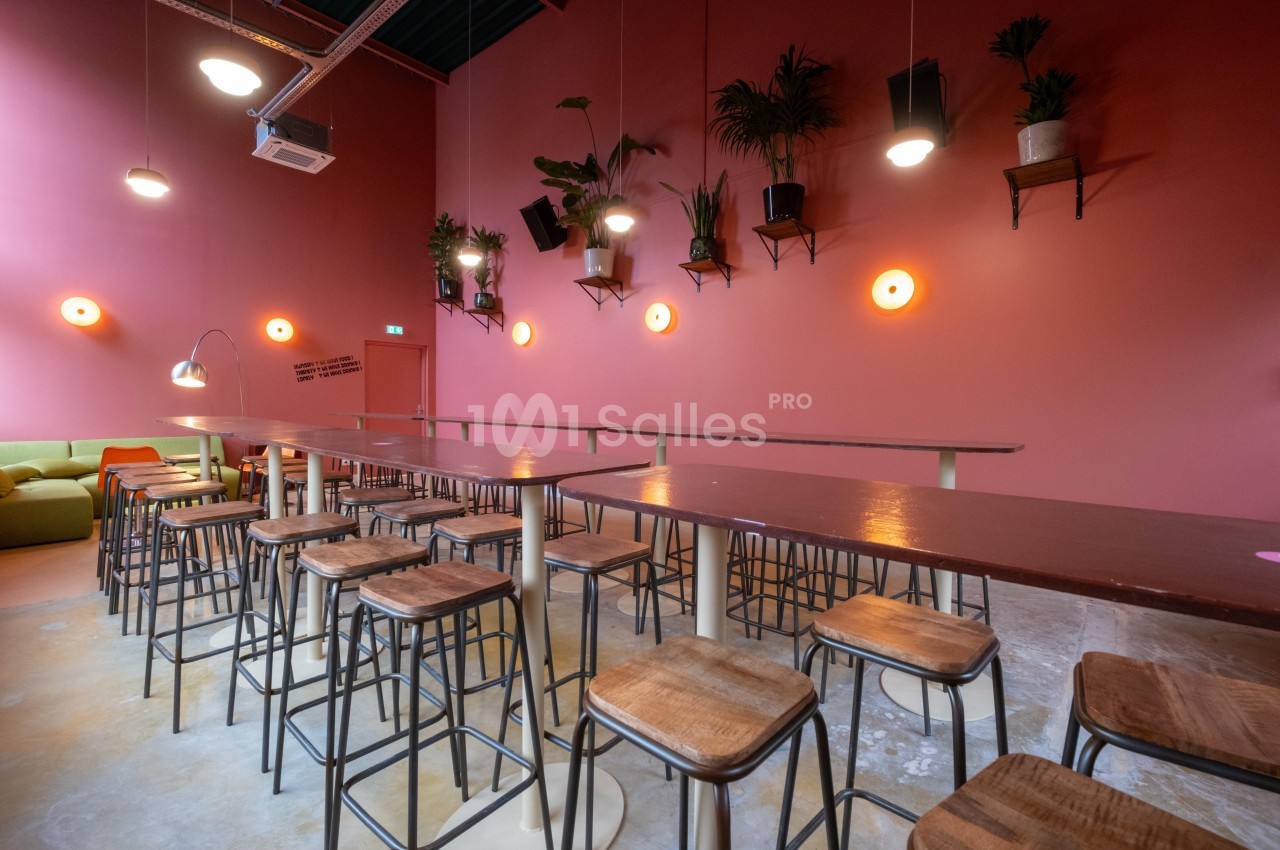 Salle avec murs roses, longues tables en bois, tabourets hauts et plantes suspendues sous un éclairage chaleureux.