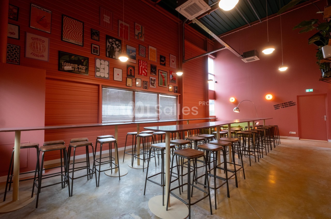 Salle lumineuse avec murs rouges, tables hautes en bois, tabourets métalliques et décorations murales colorées.