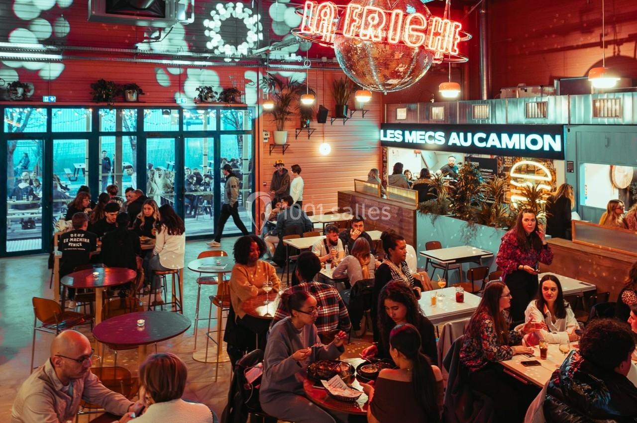 Salle animée avec des groupes de personnes assises à des tables, éclairage coloré et enseignes lumineuses au mur.