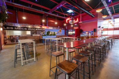 Salle lumineuse avec murs rouges, tables hautes en bois et métal, tabourets, affiches décoratives et luminaires modernes.