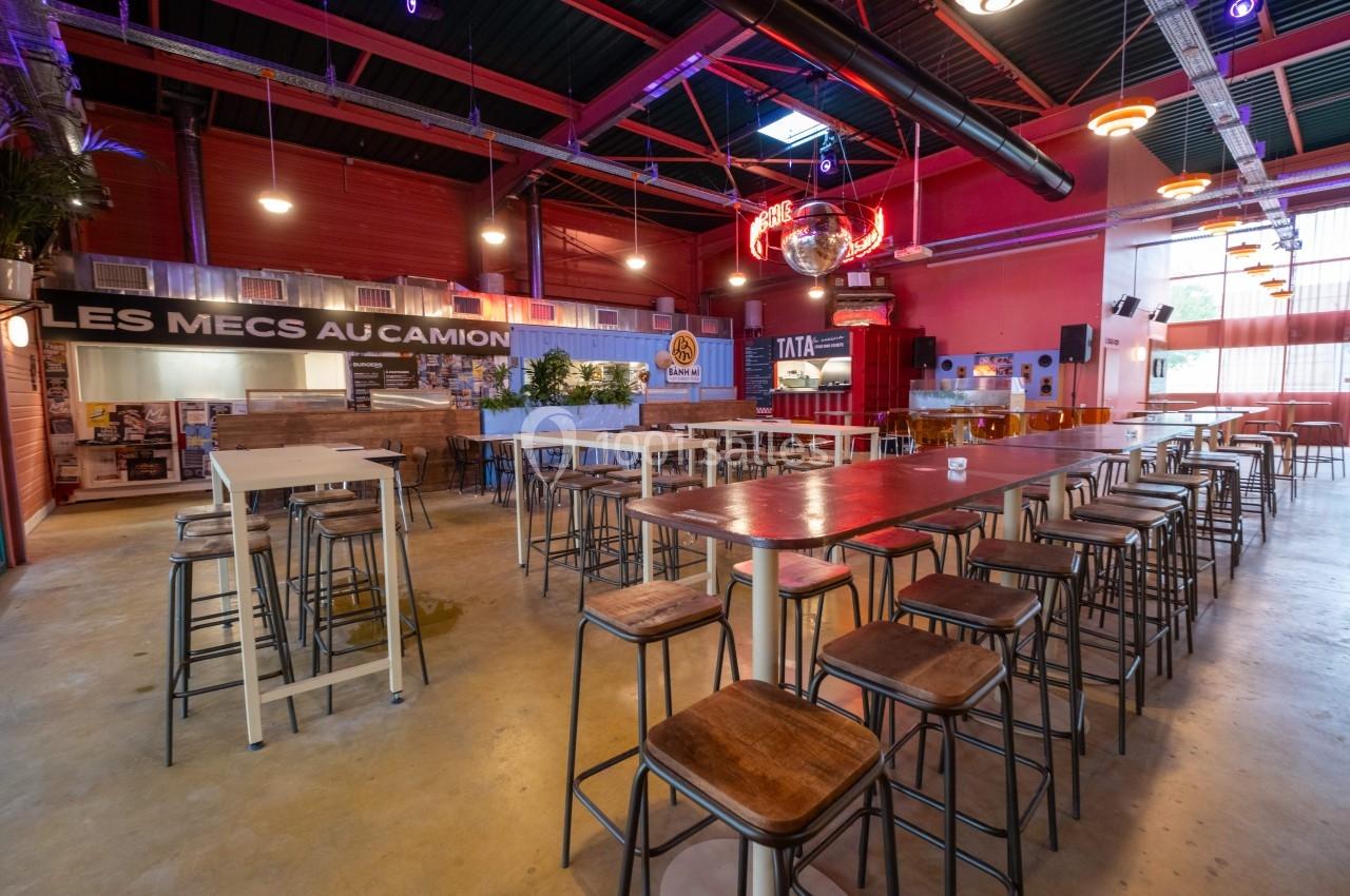 Salle spacieuse avec tables hautes, tabourets, décor industriel et enseignes lumineuses.