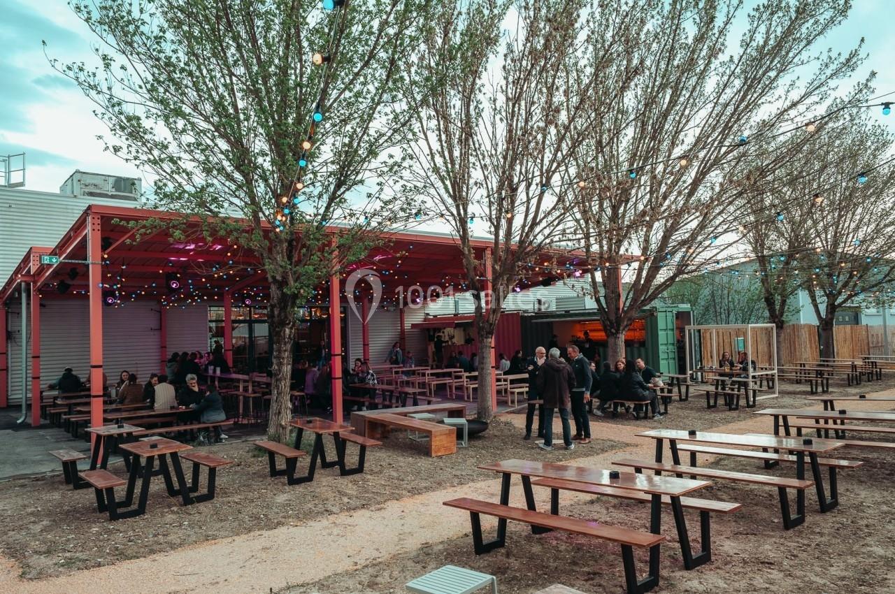 Terrasse extérieure avec tables en bois, éclairée par des guirlandes lumineuses, entourée d'arbres et de bâtiments.