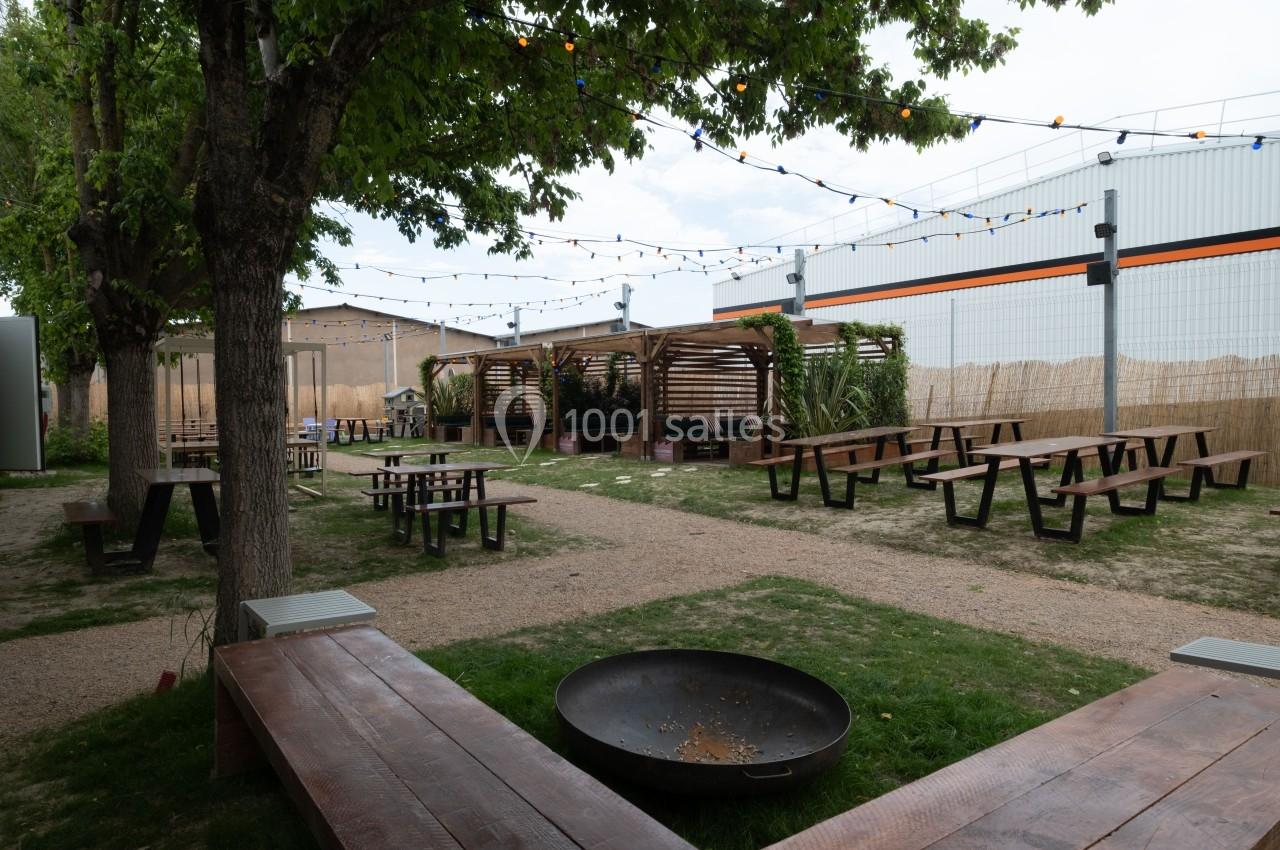 Espace extérieur avec tables de pique-nique, bancs en bois, guirlandes lumineuses et végétation sous une pergola.