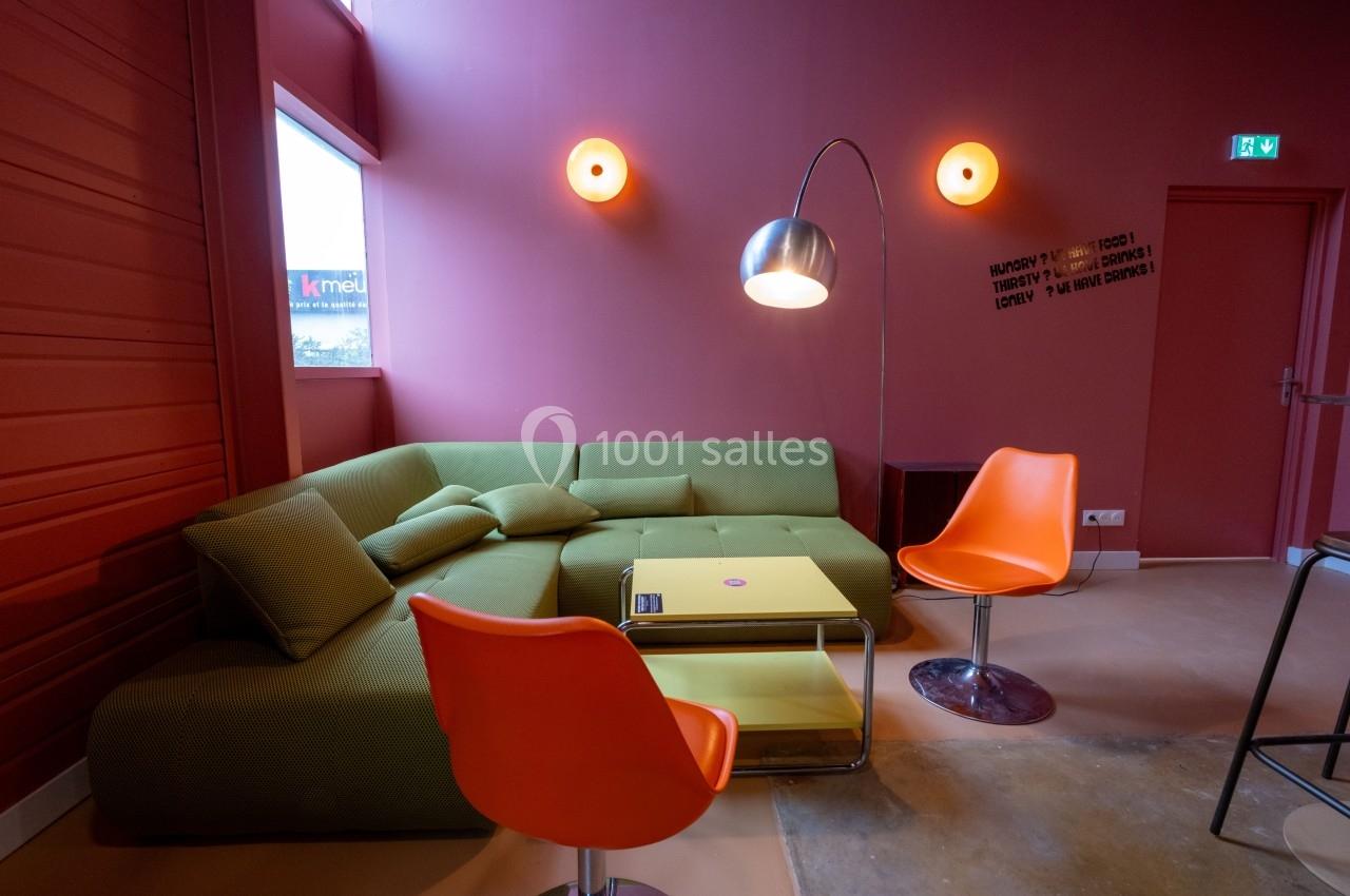 Salon moderne avec canapé vert, deux chaises orange, table basse jaune et lampe sur pied dans une pièce aux murs rouges.