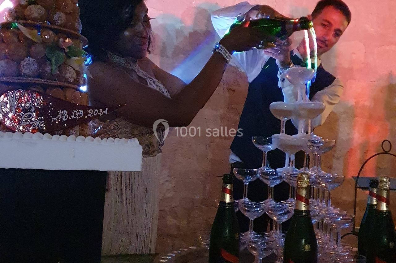 Une femme verse du champagne dans une pyramide de verres lors d'une réception festive, avec un gâteau en arrière-plan.