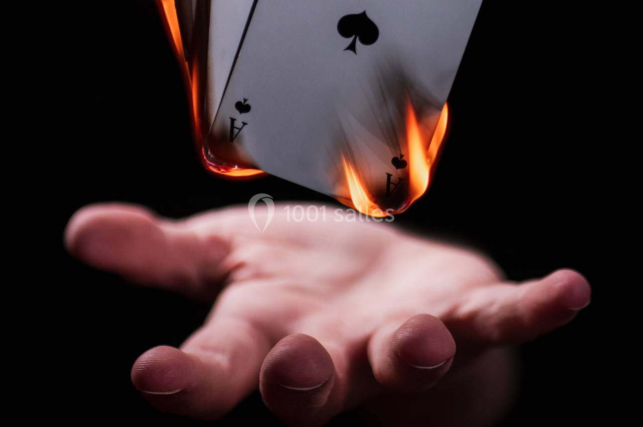 Deux cartes enflammées, un as de pique et un as de cœur, flottent au-dessus d'une main tendue sur fond noir.