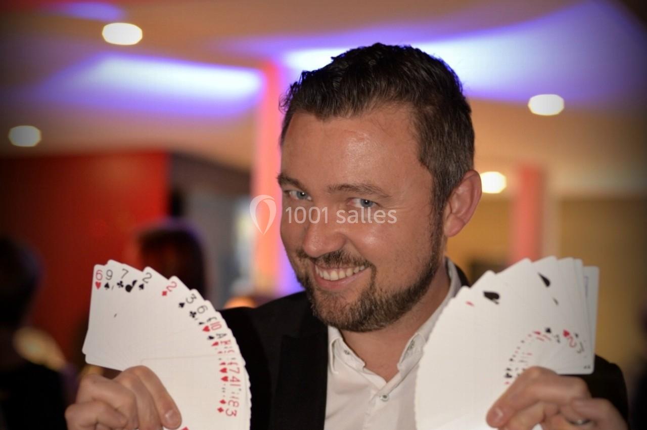 Un homme en costume souriant tient deux éventails de cartes à jouer dans un environnement lumineux.