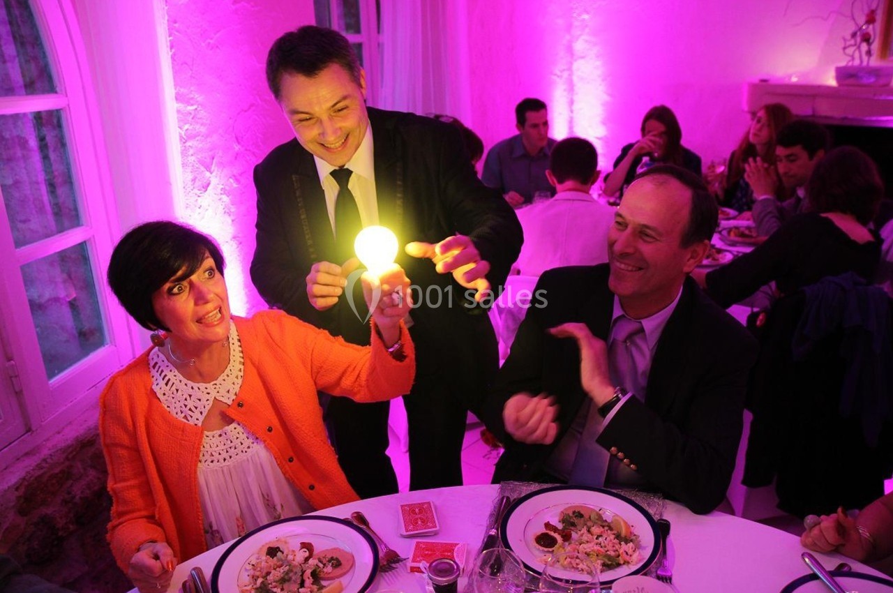 Un magicien réalise un tour avec une ampoule lumineuse devant des convives assis à table dans une ambiance festive.