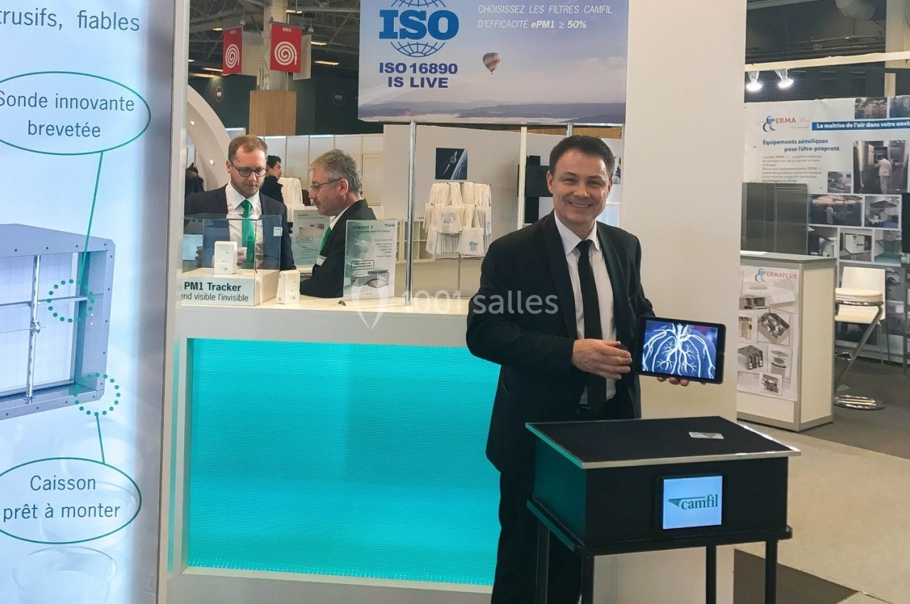 Un homme en costume présente un appareil sur un stand d'exposition, avec des affiches techniques en arrière-plan.