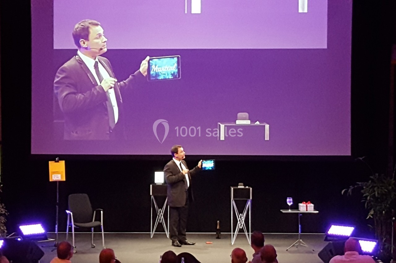 Un homme en costume présente une tablette sur scène lors d'une conférence, avec un écran projetant son image.