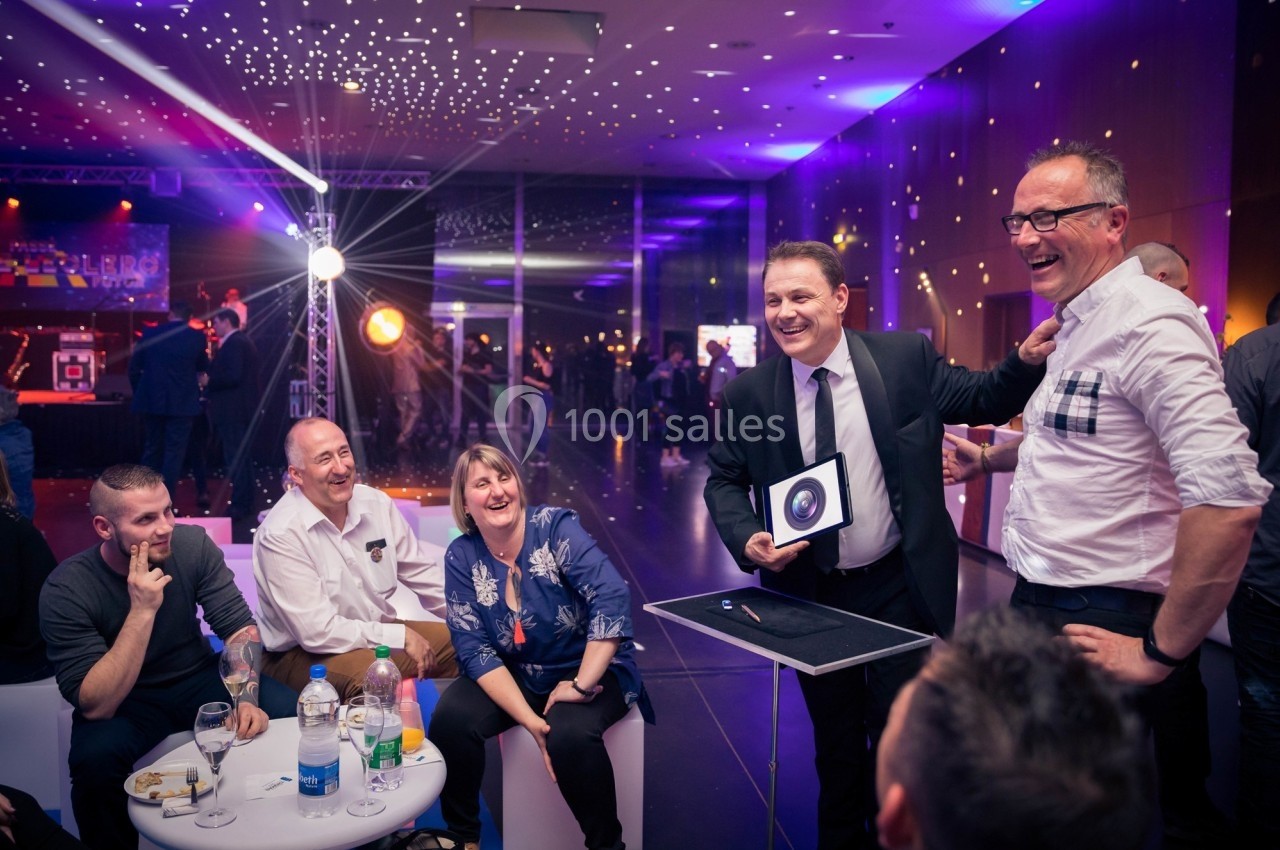 Un magicien présente un tour à un groupe de personnes souriantes dans une salle éclairée de lumières colorées.