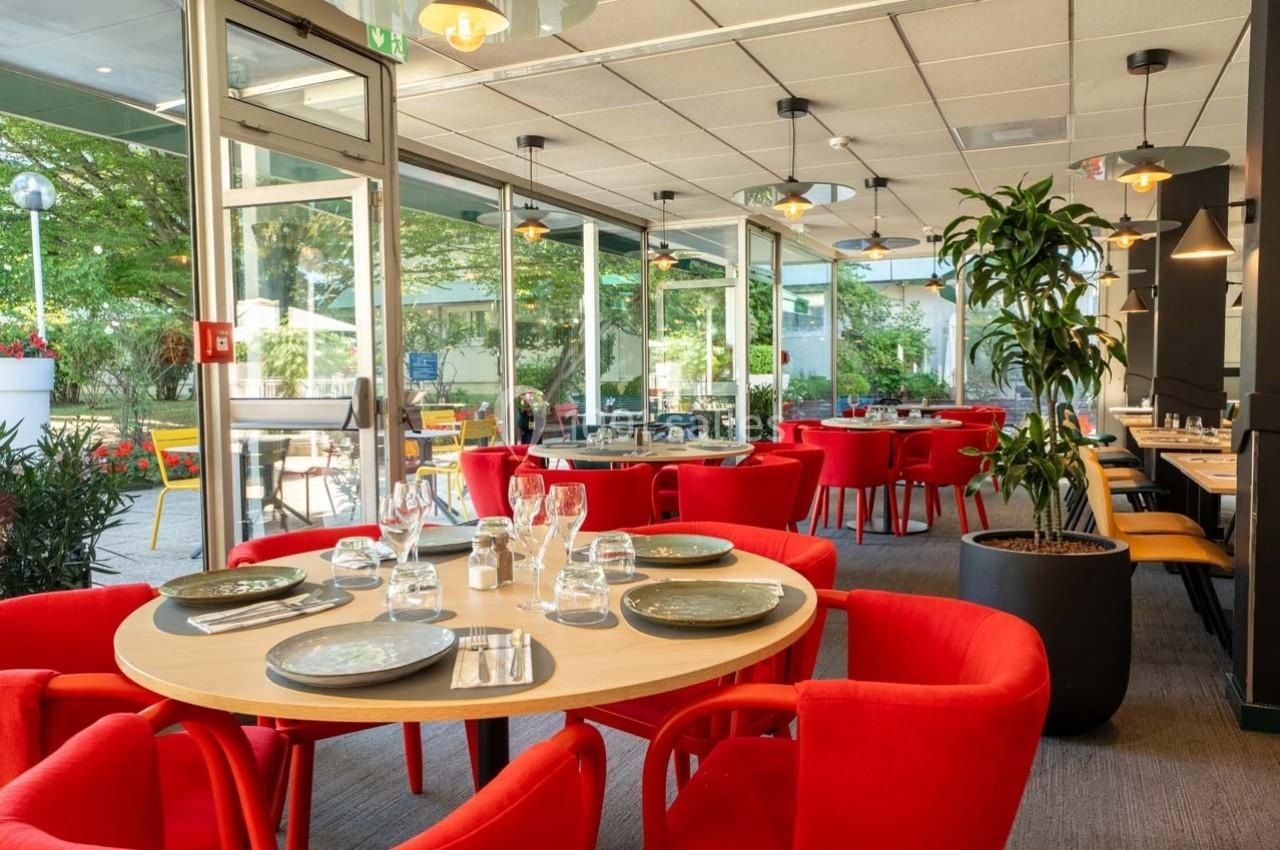 Location salle Orgeval (Yvelines) - Novotel Poissy Orgeval #5 Salle de restaurant lumineuse avec des tables dressées, chaises rouges et vue sur une terrasse arborée.