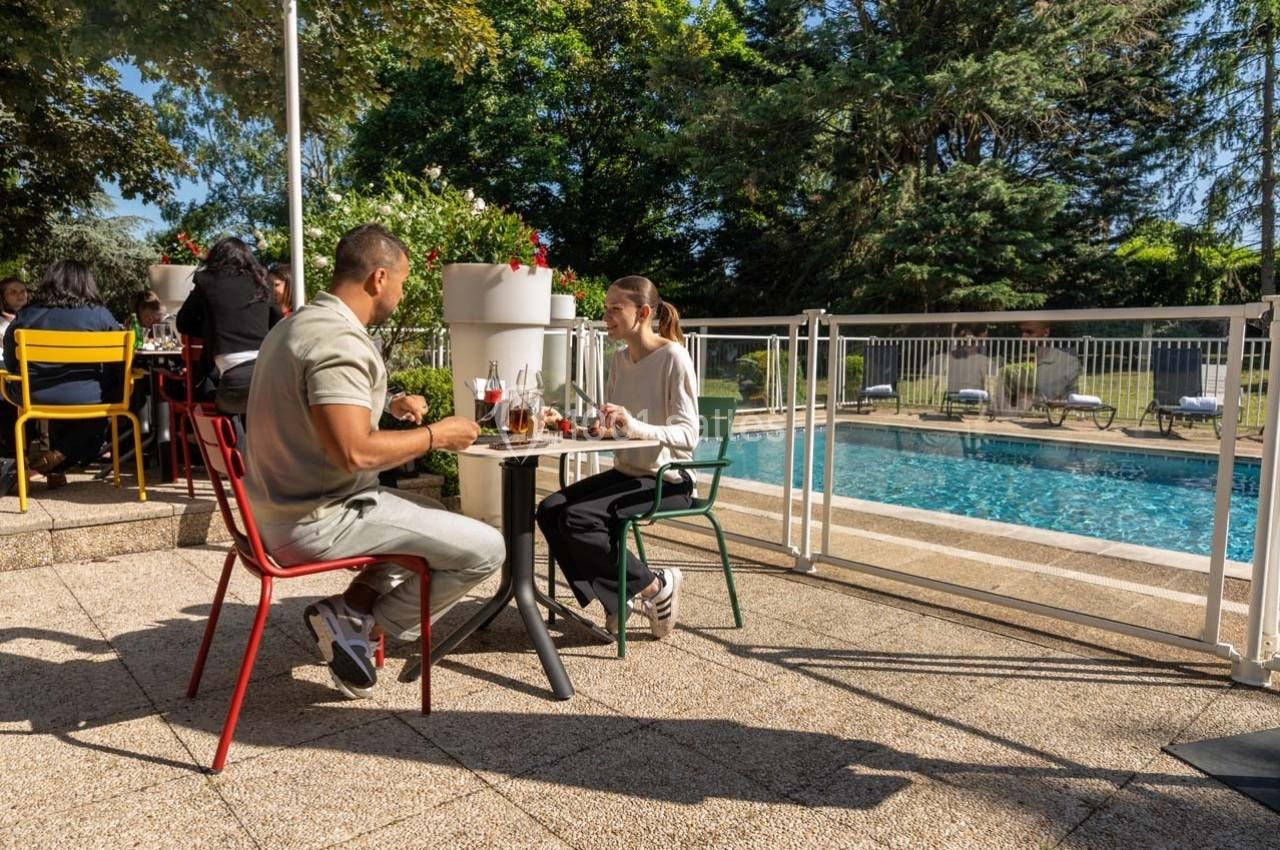 Location salle Orgeval (Yvelines) - Novotel Poissy Orgeval #20 Deux personnes déjeunent en terrasse près d'une piscine, entourées de chaises colorées et d'arbres.