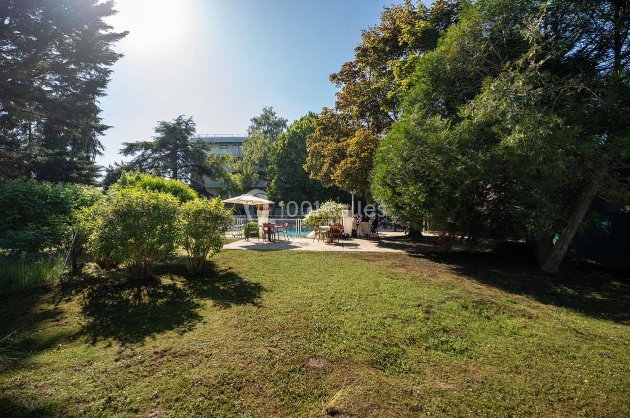 Location salle Orgeval (Yvelines) - Novotel Poissy Orgeval #21 Espace extérieur verdoyant avec pelouse, arbres et une piscine entourée de chaises et parasols sous un ciel ensoleillé.