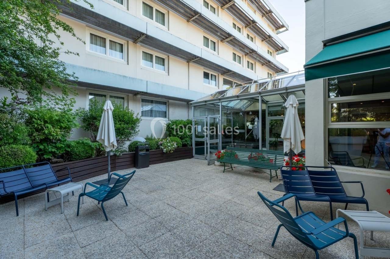 Location salle Orgeval (Yvelines) - Novotel Poissy Orgeval #26 Cour extérieure avec mobilier de jardin, parasols fermés, plantes et accès vitré à un bâtiment moderne.