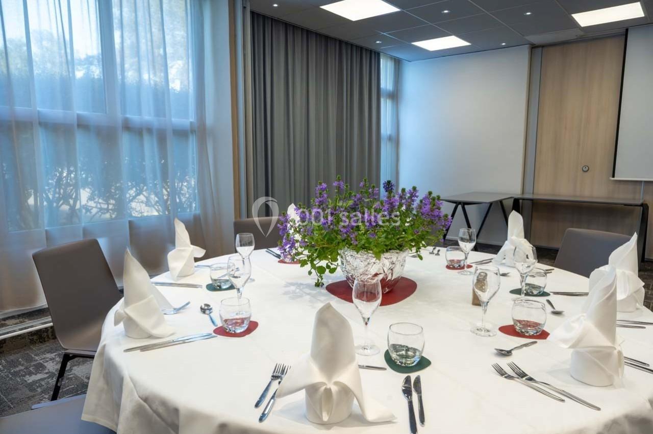 Location salle Orgeval (Yvelines) - Novotel Poissy Orgeval #3 Table ronde dressée avec nappes blanches, serviettes pliées, verres et couverts, décorée d'un centre floral violet.
