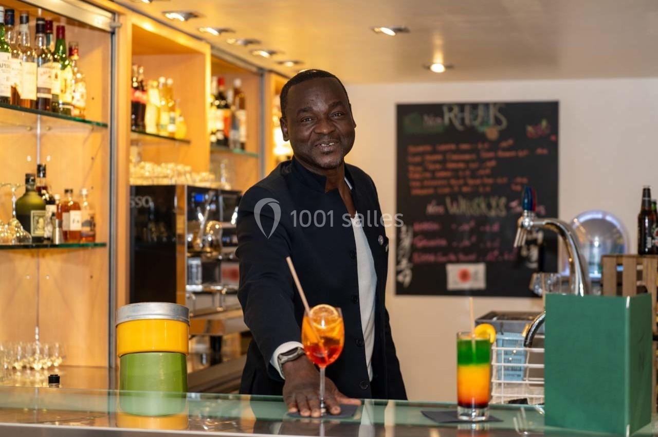 Location salle Orgeval (Yvelines) - Novotel Poissy Orgeval #28 Un barman souriant présente un cocktail coloré sur un comptoir, avec des étagères de bouteilles en arrière-plan.