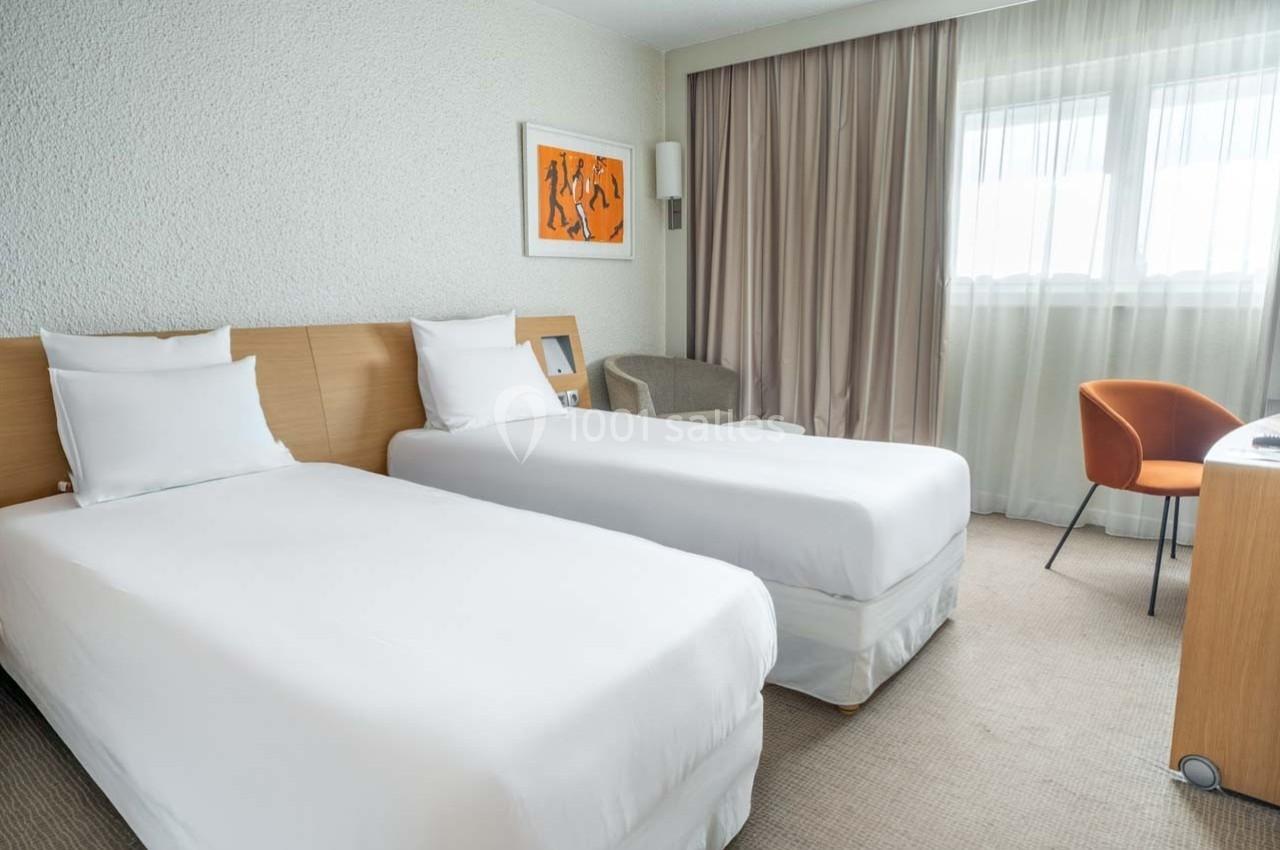 Location salle Orgeval (Yvelines) - Novotel Poissy Orgeval #34 Chambre d'hôtel avec deux lits simples, un bureau, une chaise orange et une grande fenêtre avec rideaux beige.