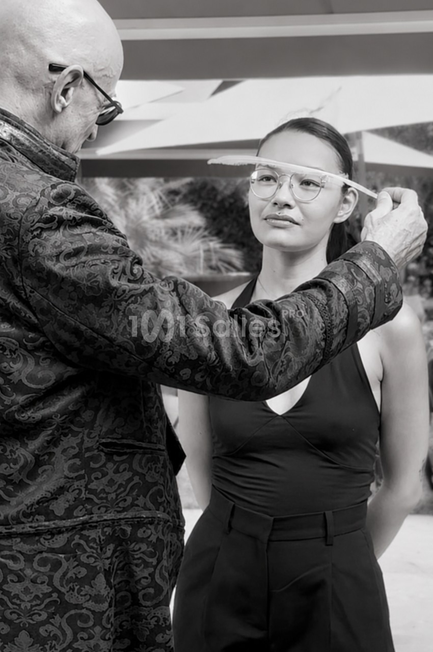 Un homme ajuste les lunettes d'une femme debout sous un auvent, dans un environnement extérieur.