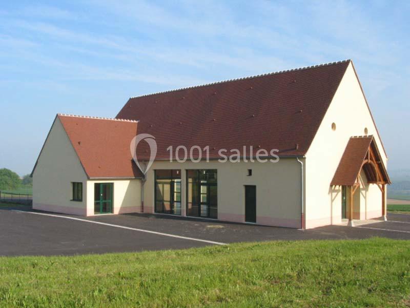 Bâtiment moderne avec toit en tuiles rouges, grandes baies vitrées et parking asphalté, entouré de verdure.