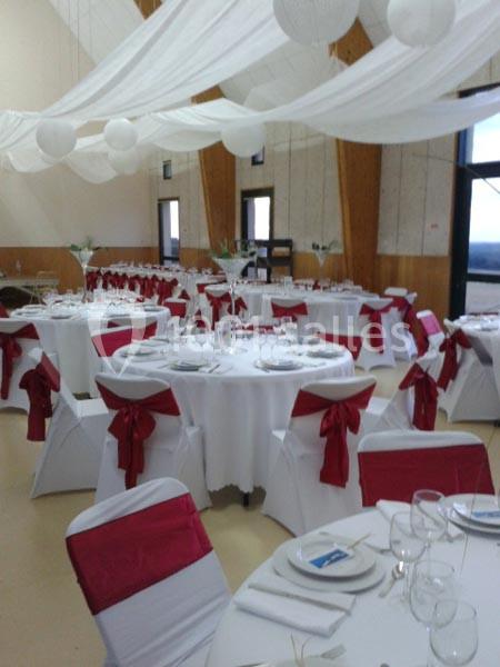 Salle de réception décorée avec des tables rondes, nappes blanches, chaises ornées de nœuds rouges et voilages suspendus.