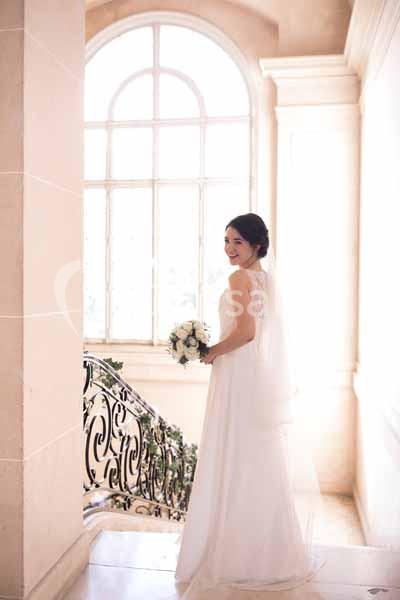 Femme en robe de mariée tenant un bouquet, debout près d'un escalier orné, devant une grande fenêtre lumineuse.