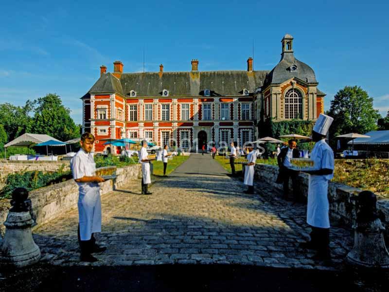 Des serveurs en tenue blanche tenant des plateaux devant un château en briques rouges sous un ciel dégagé.