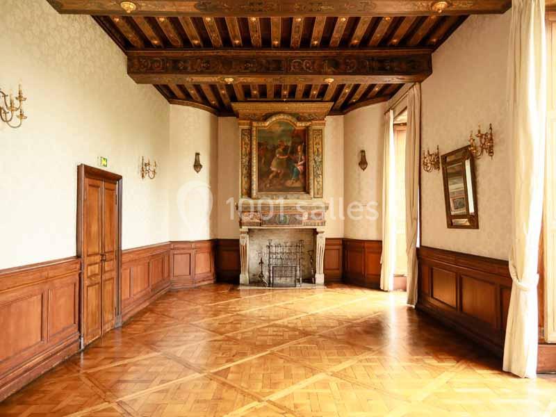 Salle lumineuse avec parquet en bois, cheminée ornée, plafond à poutres apparentes et murs décorés de boiseries.