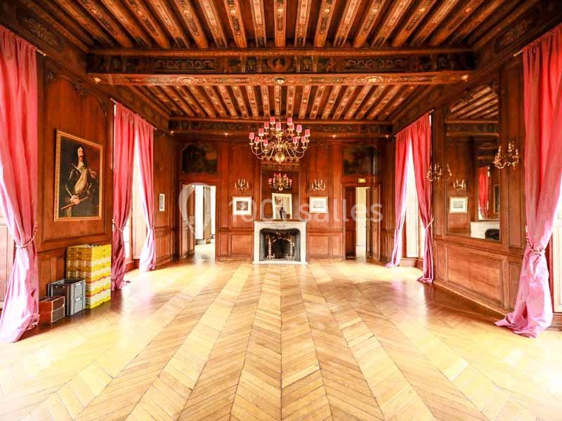 Salle avec parquet en bois, murs lambrissés, cheminée, lustres et rideaux roses encadrant de grandes fenêtres.