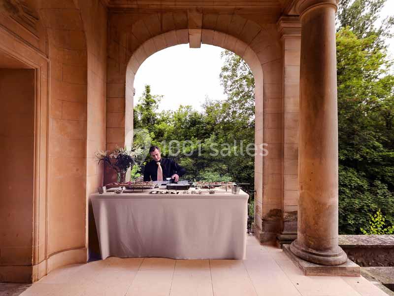 Un DJ mixe sur une table de mixage sous une arche en pierre, avec un fond de verdure.
